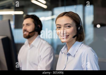 Friendly customer service representative wearing a headset and smiling at the camera, with a colleague working in the background, depicting efficient support and communication Stock Photo