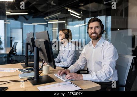 Smiling call center agent in a modern office wearing a headset and working at a computer, providing customer support while a colleague works in the background, professional teamwork Stock Photo
