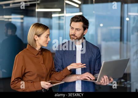 Two business professionals standing in a modern office, one man holding a laptop and the woman holding a tablet, engaging in a discussion and working together Stock Photo