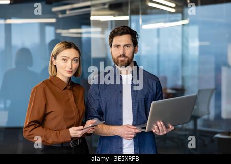 Two confident business professionals, a woman holding a tablet and a man holding a laptop, standing together in a contemporary office setting, representing teamwork and innovative collaboration Stock Photo