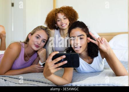 Diverse female friends lying on bed sharing smartphone in bedroom with textured blanket and pillows Stock Photo