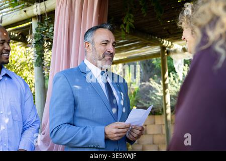 Small group standing under wooden pergola in garden, man in light blue suit reading folded paper Stock Photo