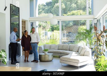 Diverse couple talking to African American agent showing glass walls and sectional sofa Stock Photo
