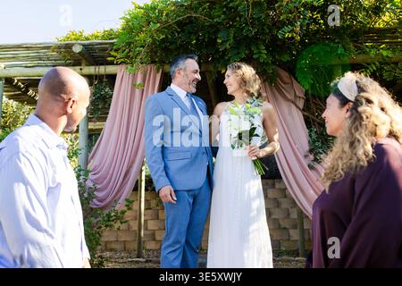 Vows starting, newlyweds exchanging under pergola draped pink, bride in white dress holding lilies Stock Photo