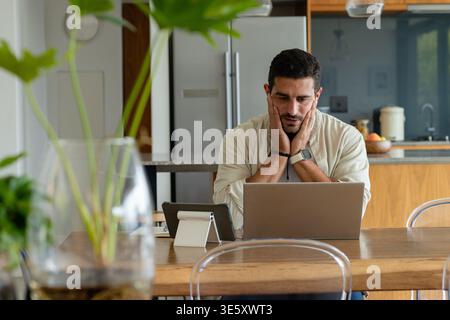 Man sitting at wooden dining table in modern kitchen working on laptop with tablet, glass vase Stock Photo