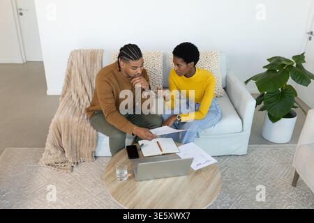 Reviewing couple sitting on sofa in living room, with laptop, printed documents and smartphone Stock Photo