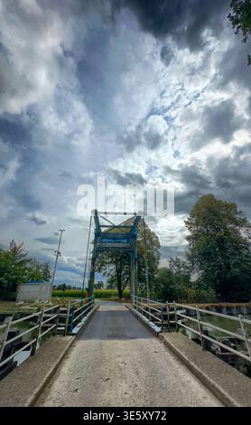 Rustic metal bridge under dramatic clouds, narrow single-lane crossing over shallow stream, wet surface with puddle reflections, Stock Photo