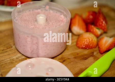 Close-up of a plastic blender bowl filled with pink strawberry smoothie. Slices of fresh ripe strawberries and a kitchen knife lie nearby on a wooden cutting board. Natural soft lighting emphasizes the texture of the food and creates an atmosphere of home-cooked healthy breakfast. of making strawberry smoothie in a blender on a wooden board, fresh chopped berries. High quality photo Stock Photo