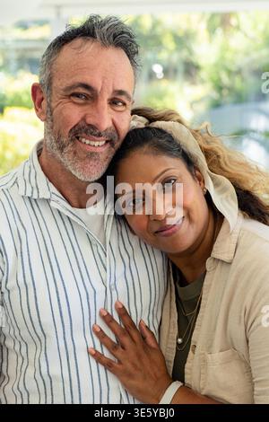Diverse couple standing close by window at home, man wearing striped shirt, woman wearing headband Stock Photo
