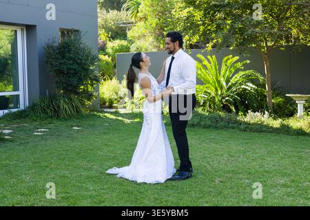 Diverse couple standing on lawn holding hands, smiling, wearing white lace dress and black necktie Stock Photo