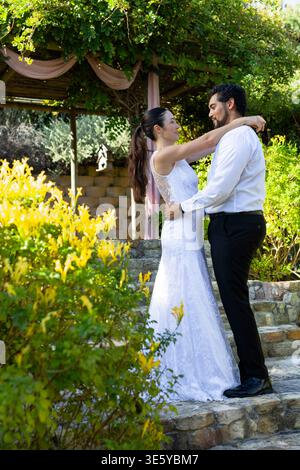 Asian bride African American groom in attire posing on stone steps under draped garden pergola Stock Photo