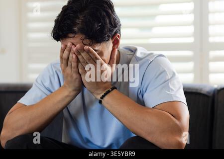 Asian man sitting on dark sofa in living room, covering face, showing black beaded bracelet Stock Photo
