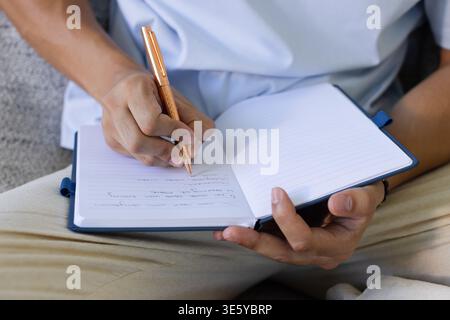 Asian adult man sitting on couch wearing white shirt writing in notebook with rose-gold pen Stock Photo