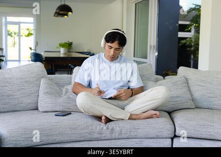 Asian man writing in spiral notebook with pen on gray sofa wearing blue-tee and headphones Stock Photo