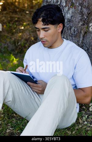 Asian man sitting on grass writing in notebook with metal pen wearing light-blue shirt, beige pants Stock Photo