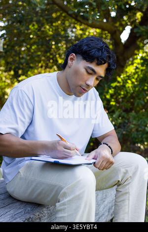 Adult male sitting on log at park writing on pad with orange-pen, white tee, beaded bracelet Stock Photo