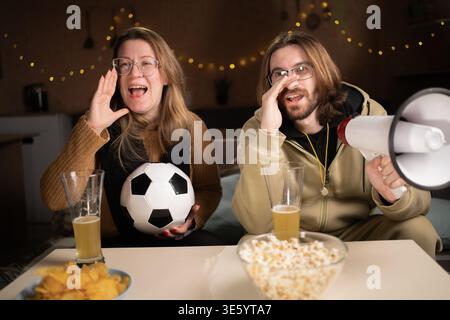 Excited emotional football fans couple with soccer ball watching football game on tv celebrating goal at home. copy space Stock Photo