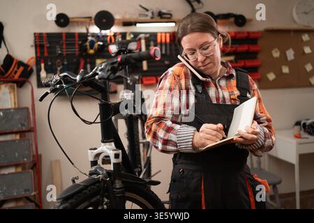 Female bike repairman repairing mountain bicycle with equipment for mechanic or technician writing notes in workshop. Bicycle repair, maintenance and Stock Photo