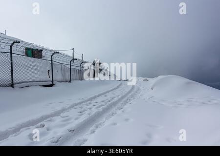 View of snow-covered ground with tire tracks leading towards a fence under a cloudy sky, Pir Chanasi, Azad Kashmir, Pakistan. Stock Photo