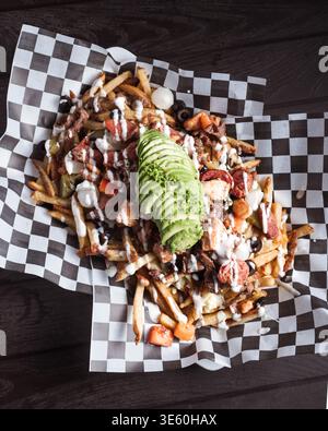 Fries piled high with toppings and avocado slices. Drizzled sauces and crispy bacon add bold texture. Checkered paper holds the dish on dark wood. Per Stock Photo