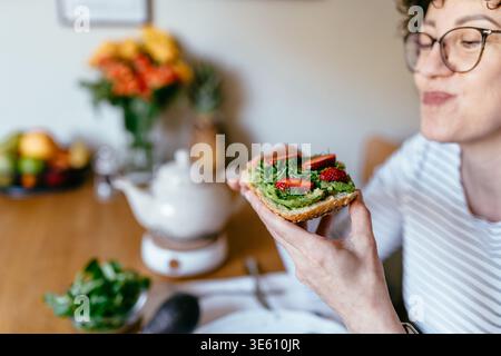 Middle aged woman holding avocado toast with strawberries close up in kitchen Stock Photo