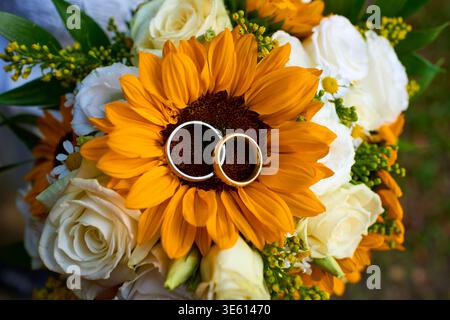 Close-up of two gold wedding rings resting on a bright yellow sunflower in a bridal bouquet with white roses. Concept of love and rustic wedding. Stock Photo