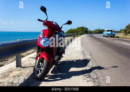 Red scooter parked on coastal road with classic vintage car in Varadero, Cuba Stock Photo
