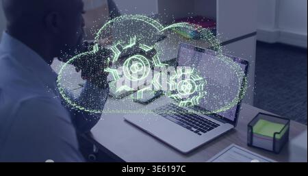 Projecting hologram with linked gears above laptop at cubicle, colleagues wearing business casual Stock Photo