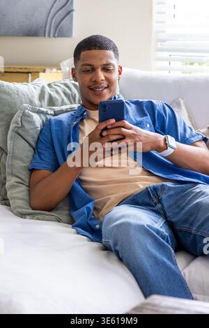African American man reclining on home sofa, in blue shirt, holding blue phone and white smartwatch Stock Photo