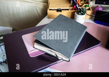 Notebooks are placed on a desk. One notebook is grey and slightly open. There are other items such as a plant and colorful pens in the background. Stock Photo