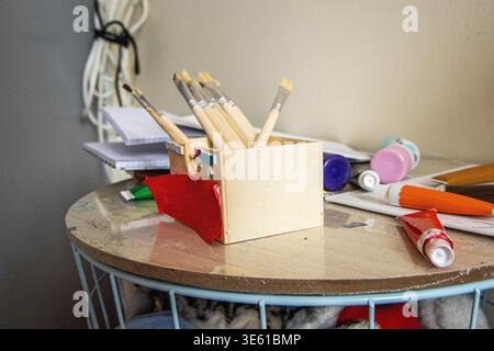 A wooden box sits on a round table. Inside the box are various pens and paintbrushes. Nearby are papers and other art supplies scattered on the table. Stock Photo