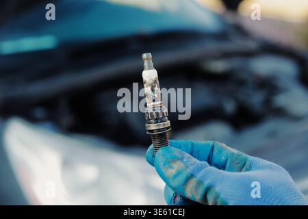 Auto mechanic holds burned spark plug in hand close up with car in background. Vehicle maintenance, engine failure and repair service concept. Stock Photo