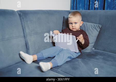 A child is seated on a couch, focused on a tablet device. The room is bright, and the child is dressed casually with socks. The child shows concentrat Stock Photo