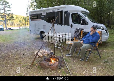 Man sitting at a small campfire in front of the motorhome, free camping on the banks of the Oesterdalaelven near Aelvdalen, Dalarnas laen, Sweden Stock Photo