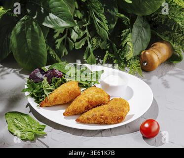 Food, Three golden-brown fried pastries are plated with fresh greens and a small bowl of dipping sauce. The setting features vibrant greenery, creatin Stock Photo