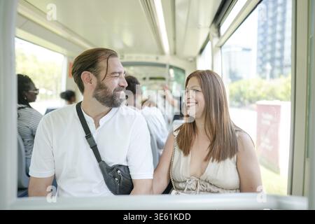 A man and woman are sitting on a bus, smiling at each other. The man is wearing a white shirt and a black backpack. The woman is wearing a white dress Stock Photo