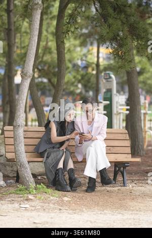 In June 2025, a Korean man and woman in their late fifties sit on a bench by the sea, chatting and smiling as they enjoy the breeze, ocean view, and c Stock Photo