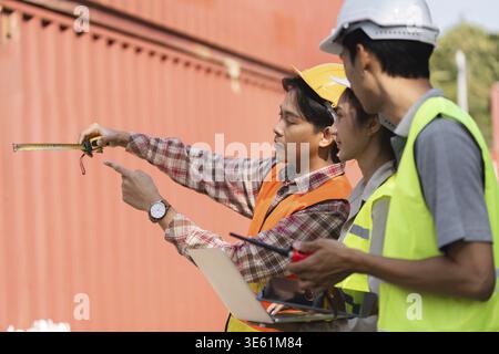 Two construction workers, an engineer and a site manager, measuring and discussing project details with blueprints at a busy construction site Stock Photo
