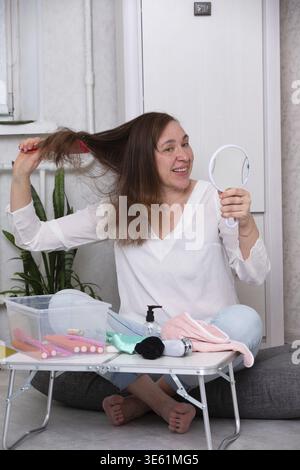 A woman is happily brushing her hair with a mirror in hand, surrounded by various hair styling tools and products in a bright, cozy indoor space Stock Photo