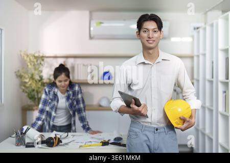 A young engineer holds a tablet and hard hat while discussing project plans with a colleague in a modern workspace Stock Photo