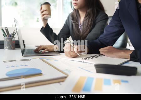 A businesswoman holds a coffee cup while taking notes on a laptop during a professional meeting focused on financial strategies Stock Photo