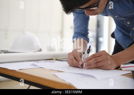 Close-up of an architect's hands using a compass to draw on house plans. The desk has design tools like a tablet, pencil, and safety helmet. Bright of Stock Photo