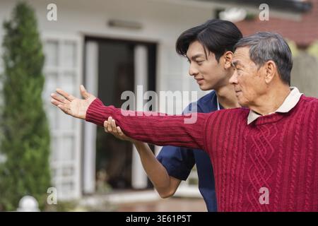 A compassionate young caregiver helps an elderly man, demonstrating a gesture of guidance in a serene home garden setting filled with greenery Stock Photo