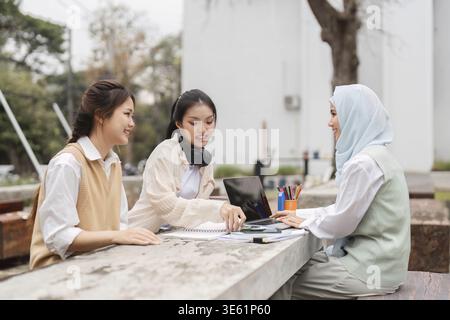 A diverse group of three women, including one in a hijab, collaborating on projects outdoors, fostering teamwork and sharing ideas in an academic envi Stock Photo