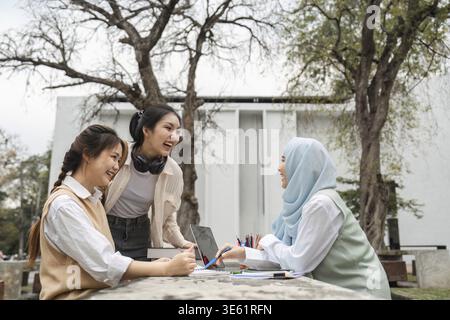 A diverse group of university students laughing and collaborating outdoors while studying, surrounded by trees and academic materials Stock Photo