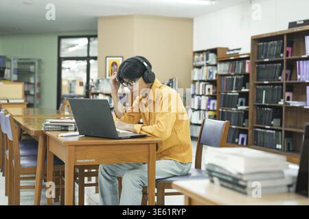 Young man student using laptop, studying, researching in library. Male student learning online at college, university. Student desk with opened book. Stock Photo