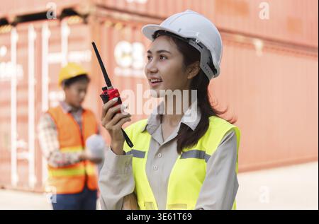 A confident female construction supervisor in a hard hat and safety vest using a walkie-talkie, coordinating with her team during an outdoor project a Stock Photo