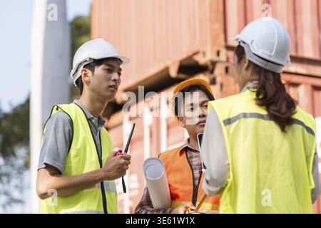 A cohesive logistics engineering team comprising a supervisor, an engineer, and a technician discussing project details and reviewing blueprints on-si Stock Photo