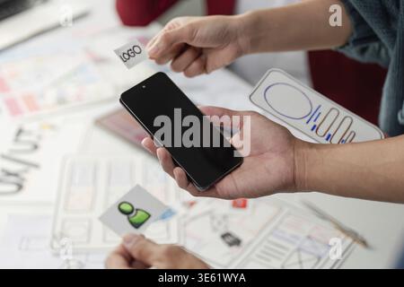 Designers hold mobile mockups and logos while discussing essential design elements to enhance user experience and create impactful branding Stock Photo