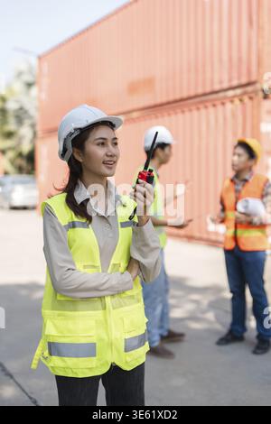 A confident female construction supervisor, wearing a hard hat and reflective safety vest, communicates over a walkie-talkie, with her colleagues enga Stock Photo